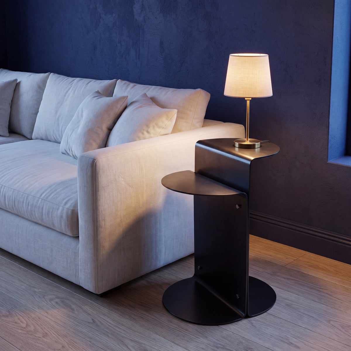 A modern black metal side table with a curved top and round base, styled in a living room next to a light grey sofa with a lit table lamp on it.