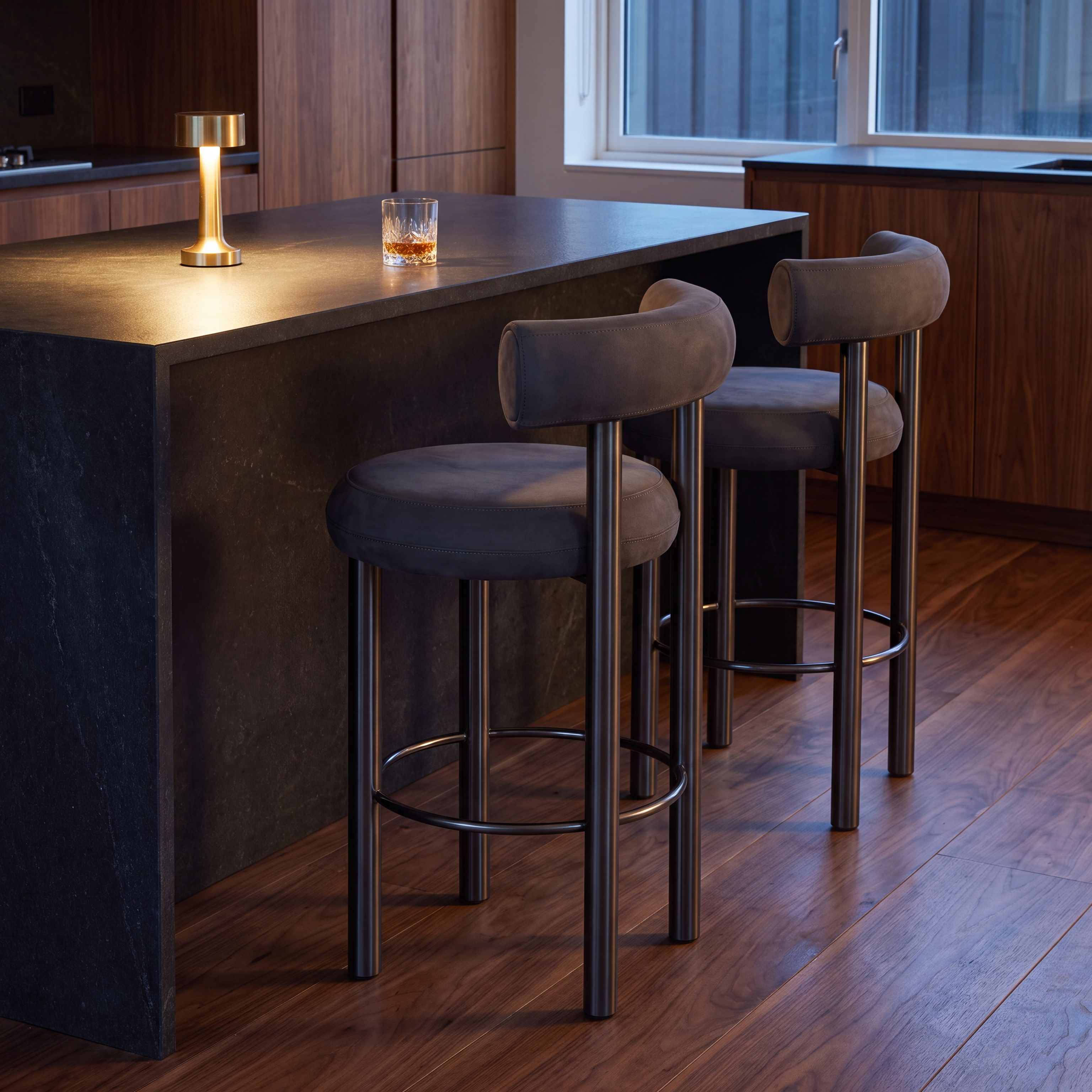 Two modern bar stools with dark gray upholstered seats and backs, black metal legs, and circular footrests, positioned at a dark stone kitchen island in a contemporary kitchen setting.