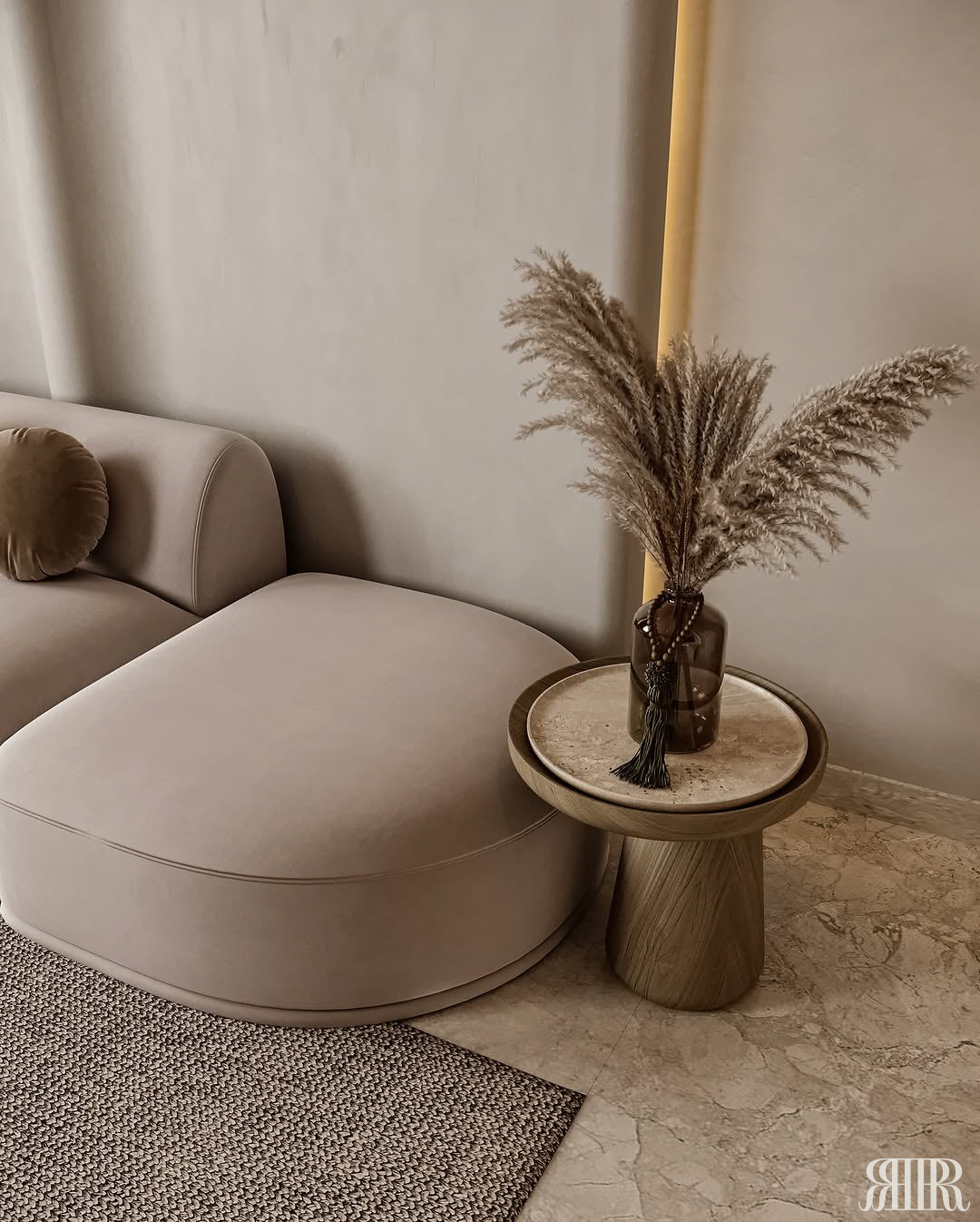 A modern beige upholstered lounge chair beside a round wooden side table holding a vase with dried pampas grass, set in a neutral-toned living room.