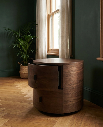 A dark brown wooden cylindrical side table with two drawers, styled in a room with dark green walls, a wood floor, and a window with sheer curtains.