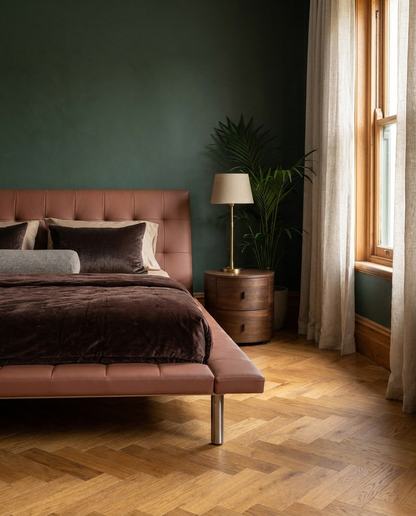 A modern bedroom scene featuring a low-profile platform bed with a tufted dusty rose leather headboard and matching footboard, styled with dark brown and gray bedding. The bed is set against a dark green wall, next to a wooden nightstand with a table lamp and a potted plant, with a window and cream curtains on the right.