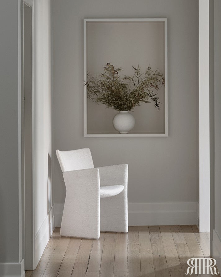 White chair in a room with a framed picture of a vase with branches on the wall.