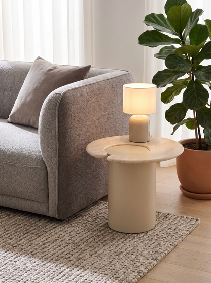 A modern side table with a cylindrical cream base and a round top, styled in a living room next to a gray sofa and a potted fiddle-leaf fig plant. A small table lamp sits on top of the table, which is placed on a woven rug over light wood flooring.