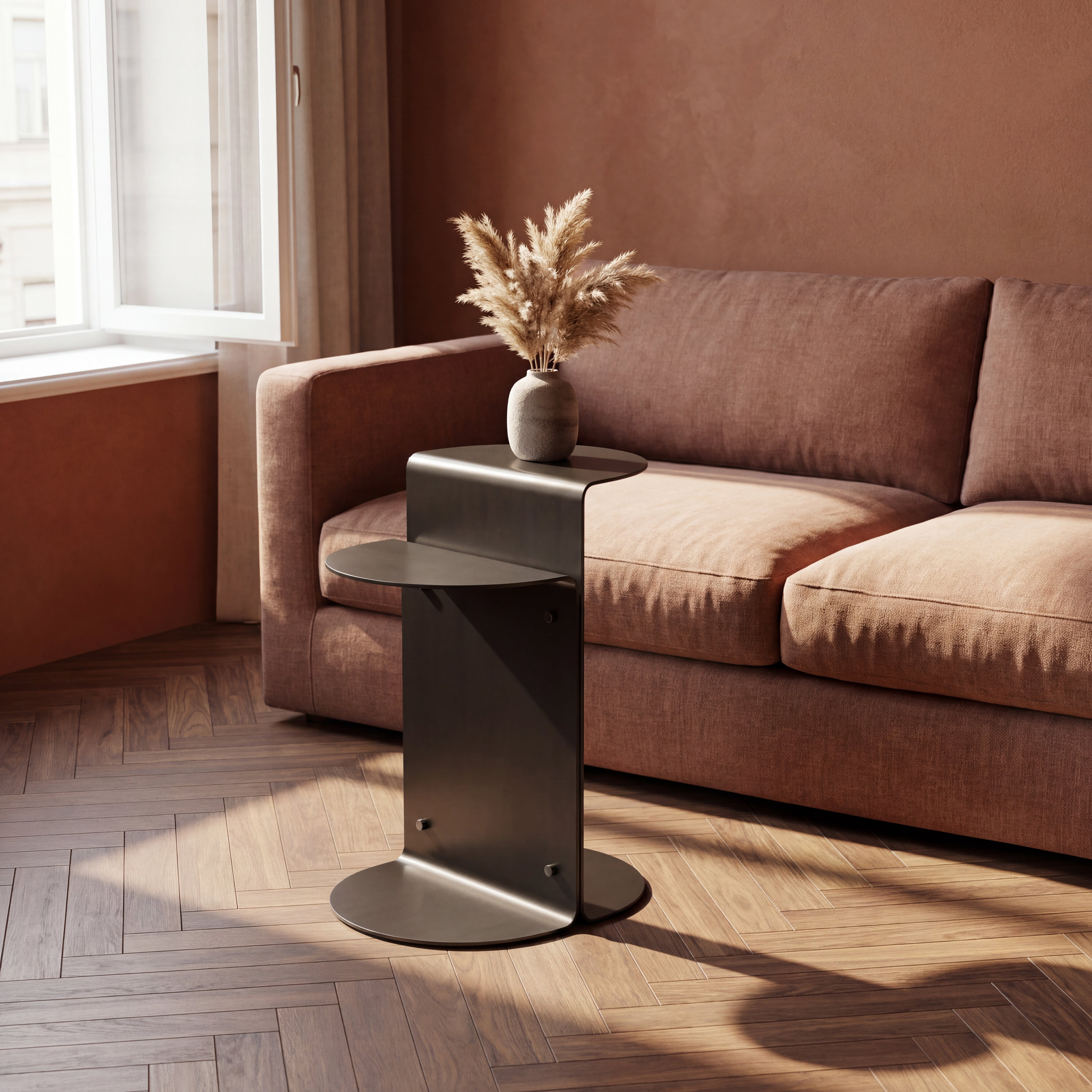 A modern dark grey metal side table with a circular base and a tiered shelf, styled in a living room next to a brown fabric sofa with a vase of dried grass.