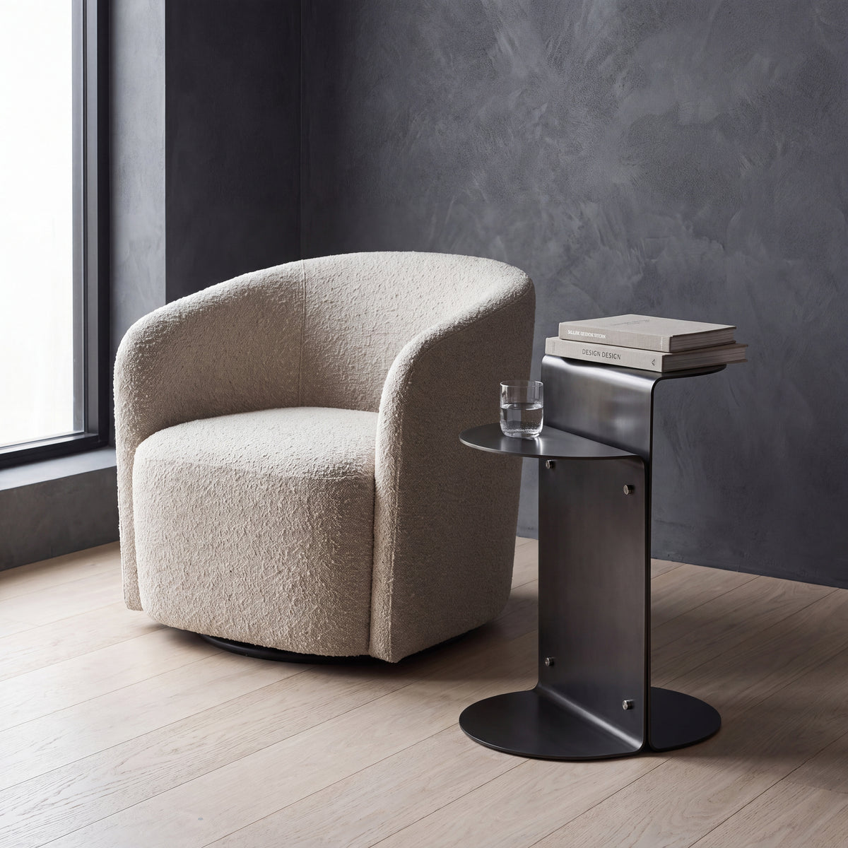 A modern beige bouclé armchair sits besides a dark metal side table with stacked books and a glass, set in a room with a dark textured wall and light wood floor. 