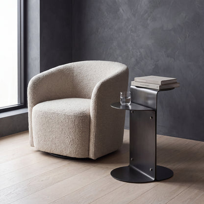 A modern beige bouclé armchair sits besides a dark metal side table with stacked books and a glass, set in a room with a dark textured wall and light wood floor. 