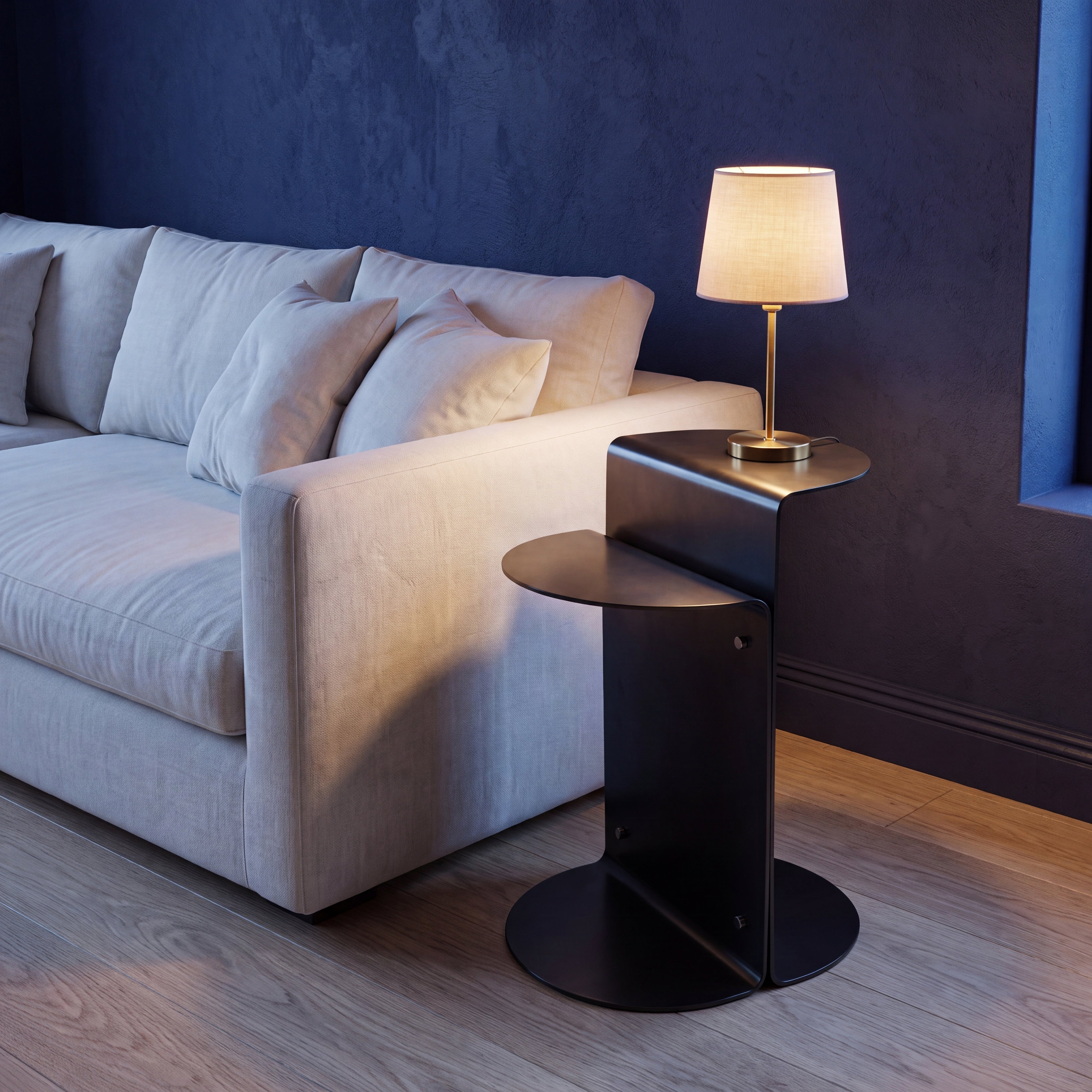 A modern black metal side table with a curved top and round base, styled in a living room next to a light grey sofa with a lit table lamp on it.