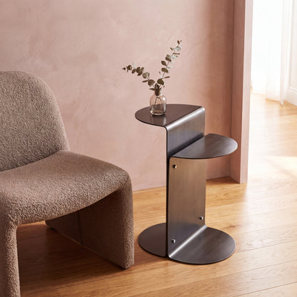 A modern sculptural side table with a dark metal circular base and two nested curved shelves, styled next to a textured brown armchair in a warm, minimalist room with a pink wall and wood floor.