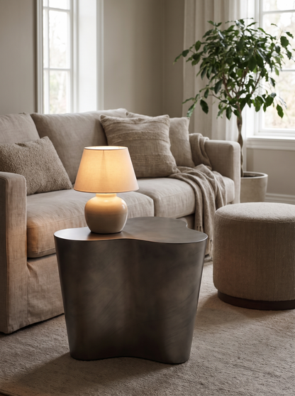 A modern wavy dark grey side table in a cozy living room setting, styled with a lit beige table lamp and surrounded by a beige sofa, textured pillows, a throw blanket, and a matching beige ottoman.