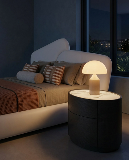 A modern mushroom-shaped table lamp with a glowing illuminated white cap and a beige base, placed on a dark round nightstand next to a bed in a dimly lit bedroom.