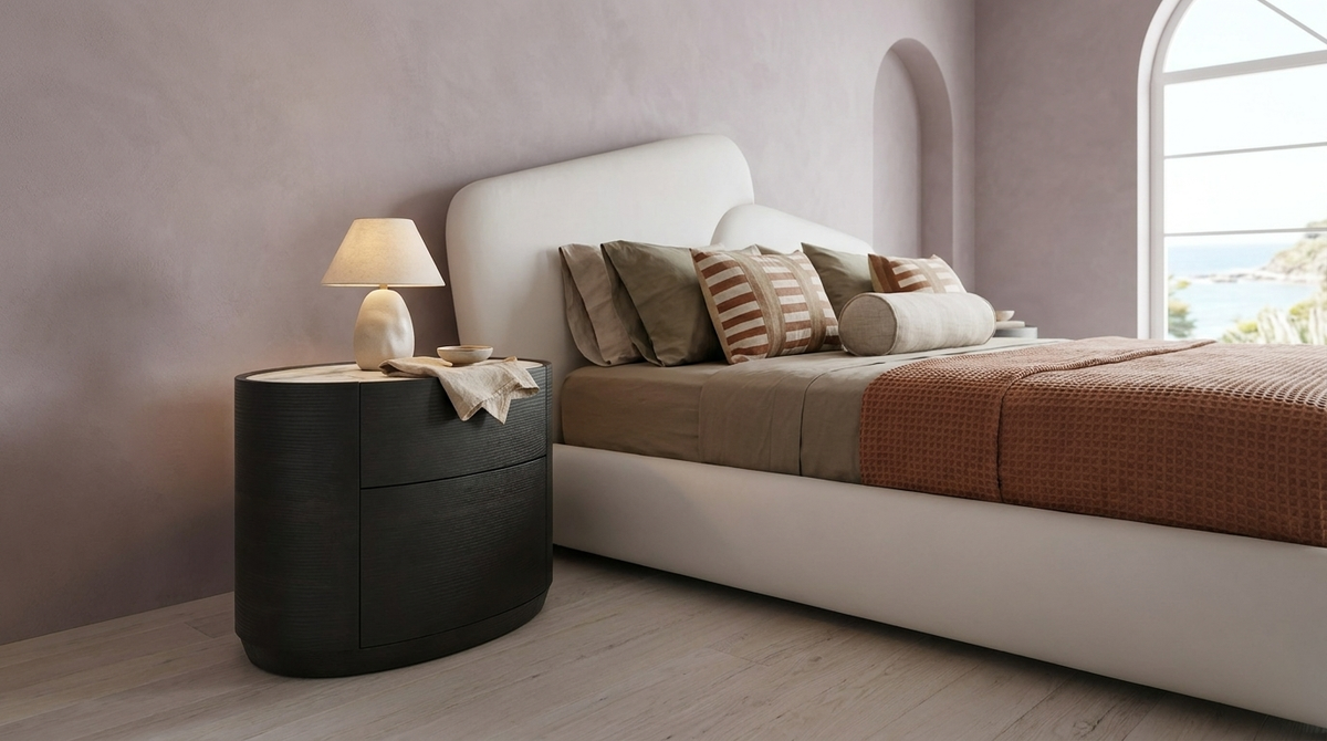 A modern bedroom scene featuring a low-profile platform bed with a white upholstered frame, styled with neutral-toned pillows and a rust-orange textured throw blanket. A dark charcoal cylindrical nightstand with a lit table lamp sits beside the bed against a textured mauve-pink wall.
