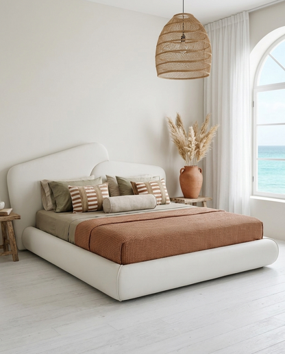 A modern platform bed with a white upholstered frame, styled with a rust-brown textured duvet, striped and solid pillows, and a woven rattan pendant light hanging above in a bright, minimalist bedroom with an ocean view.