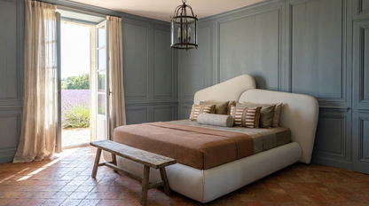 A modern upholstered bed with a curved headboard in off-white, styled with a brown duvet and striped pillows, set in a room with gray paneled walls and a rustic wooden bench at the foot of the bed.