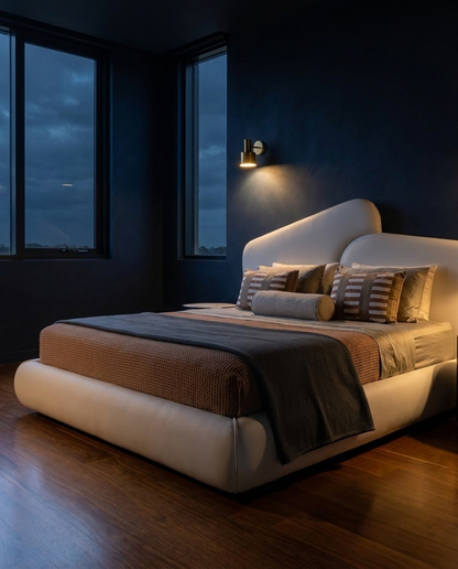 A modern low-profile platform bed with a curved headboard in light beige leather, styled with brown textured bedding and striped pillows, set in a dark navy blue bedroom with wood flooring and a wall-mounted sconce.