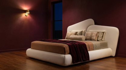 A modern low-profile platform bed with a curved, upholstered headboard and footboard in off-white leather, styled with beige and brown bedding and a dark red throw blanket, set against a dark burgundy wall with a wall-mounted sconce.
