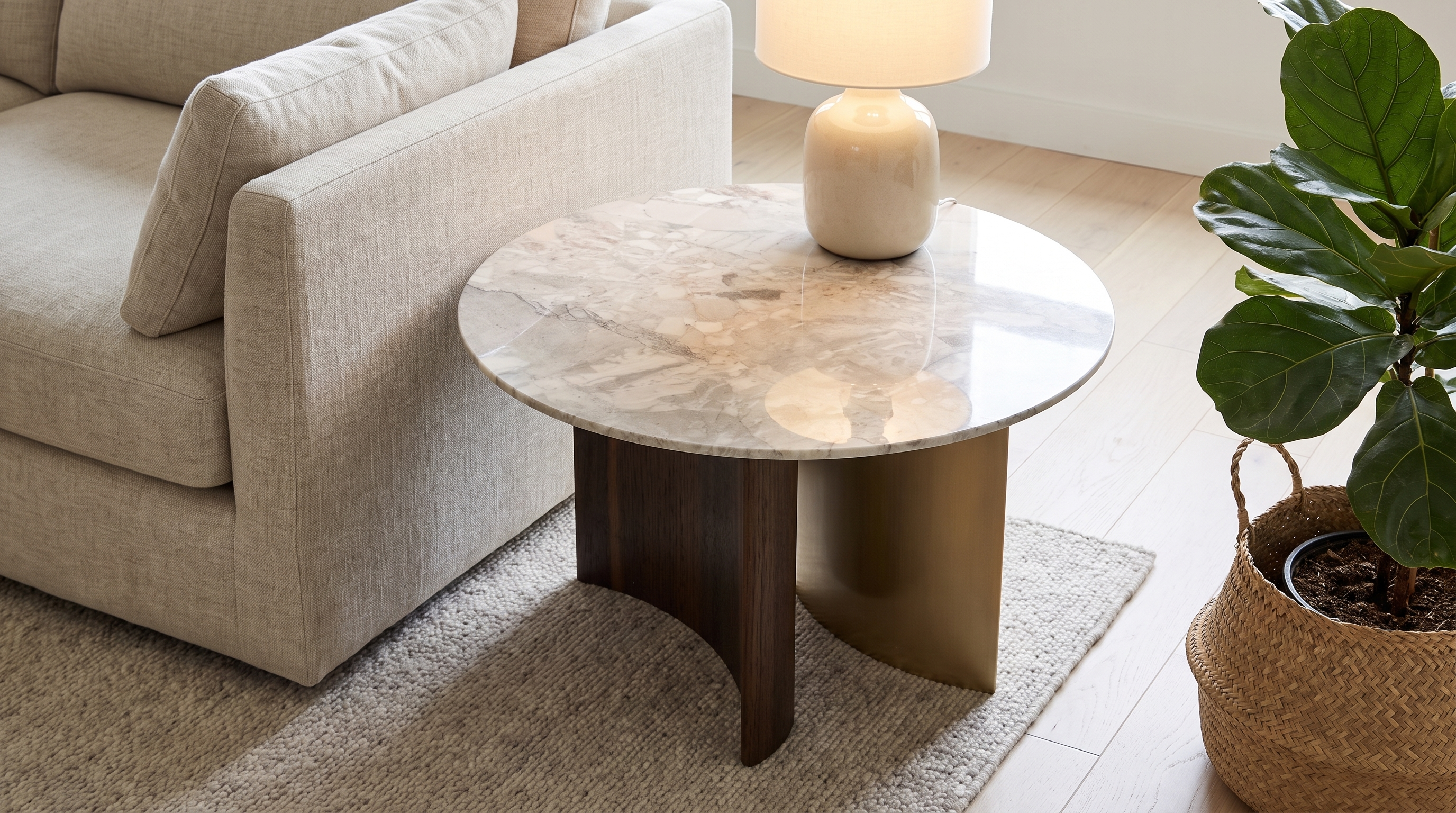 A modern round coffee table with a polished marble top and a dual-tone base of dark wood and brushed gold metal, styled in a living room next to a beige sofa and a potted fiddle-leaf fig plant.