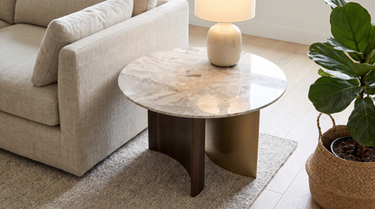 A modern round coffee table with a polished marble top and a dual-tone base of dark wood and brushed gold metal, styled in a living room next to a beige sofa and a potted fiddle-leaf fig plant.