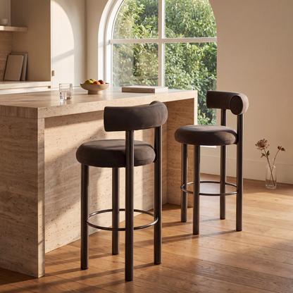 Two upholstered bar stools with dark brown frames and circular footrests, placed beside a light stone kitchen island in a sunlit room with wood floors and a large arched window.