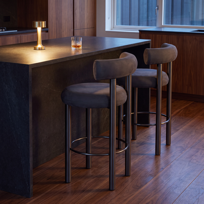 Two modern bar stools with dark gray upholstered seats and backs, black metal legs, and circular footrests, positioned at a dark stone kitchen island in a contemporary kitchen setting.