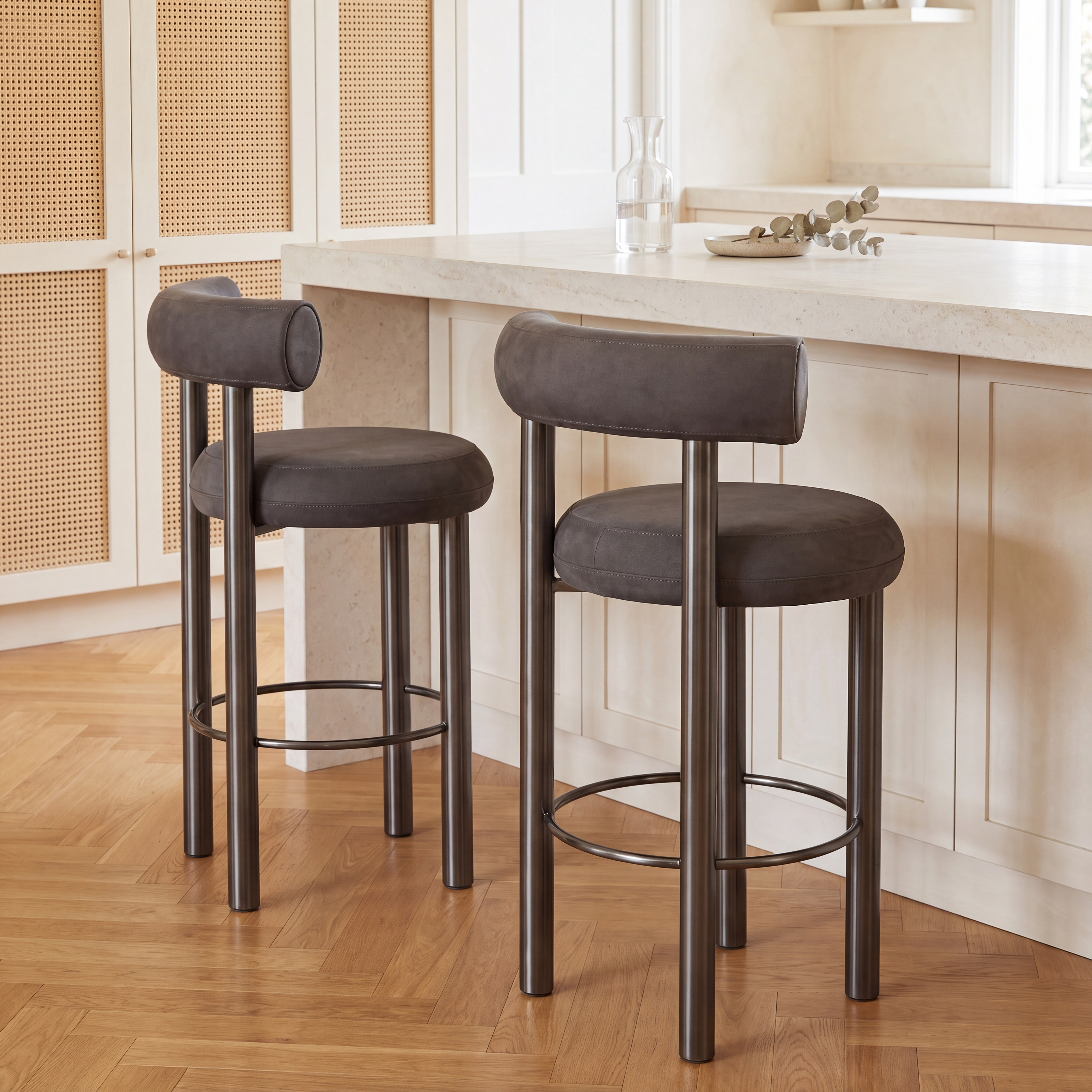 Two dark grey upholstered bar stools with round backs and dark metal legs, shown in a bright kitchen setting with a marble countertop and wood flooring.
