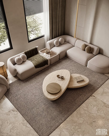 A modern living room featuring a curved modular sofa in light beige, a matching low coffee table with organic-shaped wooden tops, and a large textured brown area rug on a stone floor.