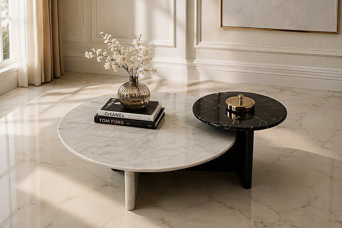 Two round marble coffee tables in a bright, elegant room. One table is white marble with a light gray veining pattern and a tapered leg, styled with books and a vase of white flowers.