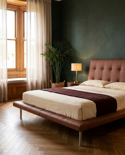 A modern platform bed with a tufted brown leather headboard and matching footboard, styled in a bedroom with a dark green wall, wood floors, a window with sheer curtains, and a bedside lamp.