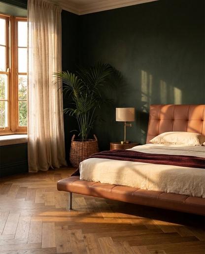 A modern bedroom featuring a low tufted brown leather bed frame with cream bedding and a dark red throw, set against a dark green wall with a potted palm and a window with warm sunlight.