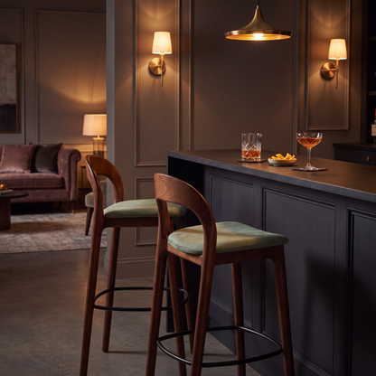 Two wooden bar stools with light green cushions sit at a dark gray kitchen island in a dimly lit, elegant kitchen. The background features a dark brown paneled wall, warm wall sconces, and a cozy living room area in the distance.