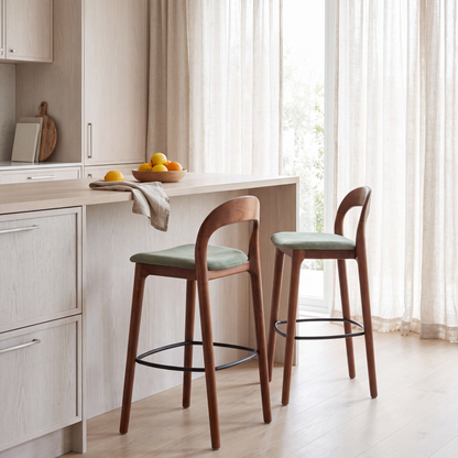 Two wooden bar stools with light green upholstered seats are placed at a kitchen island in a bright, modern kitchen with light wood cabinetry and sheer curtains.